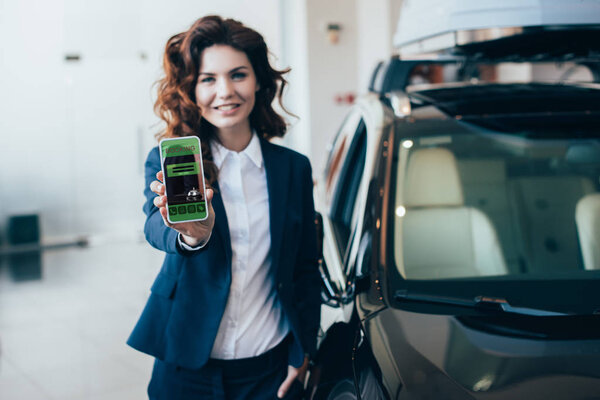 selective focus of  smiling businesswoman holding smartphone with booking app on screen and holding hand in pocket