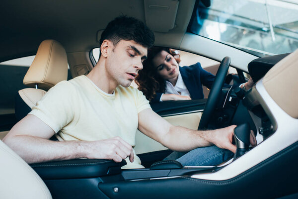 selective focus of attentive man sitting in car and pretty car dealer standing by car window