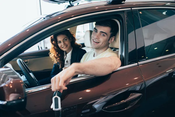 快乐的人拿着车钥匙和微笑的汽车经销商坐在新车和看着相机的选择性焦点 — 图库照片