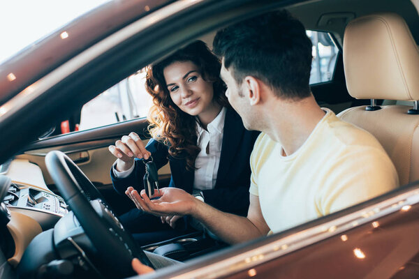 selective focus of smiling car dealer and customer sitting in new car and holding car keys