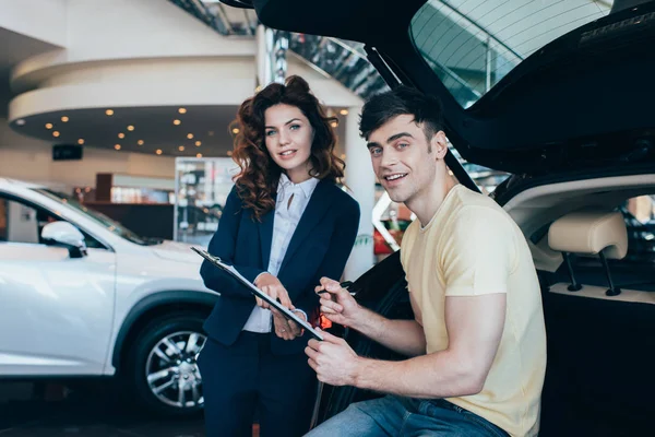 Knappe Man Ondertekening Papier Buurt Van Glimlachende Auto Dealer Kijken — Stockfoto