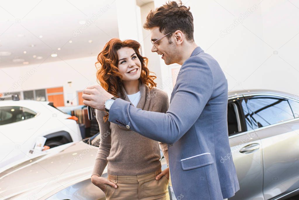 happy man in glasses looking at cheerful curly woman standing with hands in pockets in car showroom 