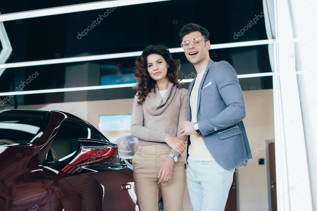 happy woman and handsome man looking at camera while standing near red automobile