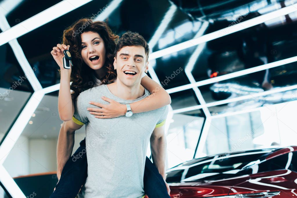 happy man piggybacking surprised curly woman with keys in hand while standing near automobile 