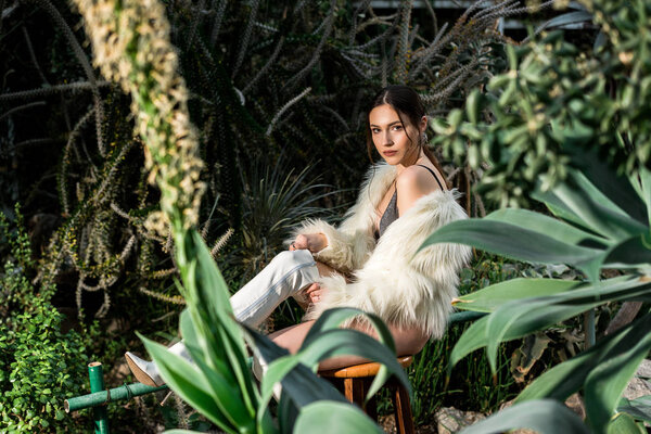 Sexy young woman in white faux fur coat sitting on chair in botanical garden