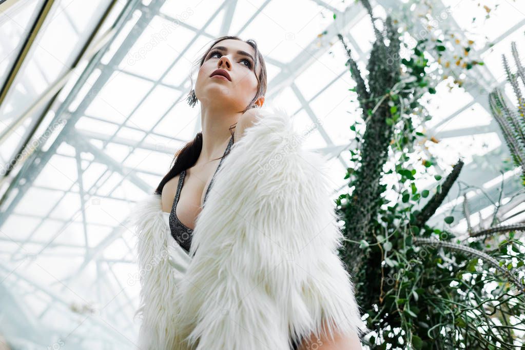 Low angle view of young woman in faux fur coat looking away in orangery