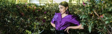 Panoramic shot of elegant young woman in purple blouse standing with arms akimbo in orangery clipart