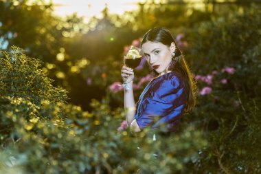 Stunning young woman in purple blouse holding wine glass in botanical garden clipart
