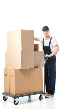 handsome mover in uniform smiling while transporting cardboard boxes on hand truck isolated on white clipart