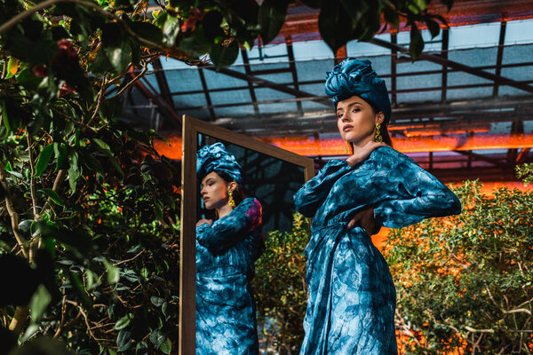 Beautiful woman in blue turban and dress standing near mirror in orangery