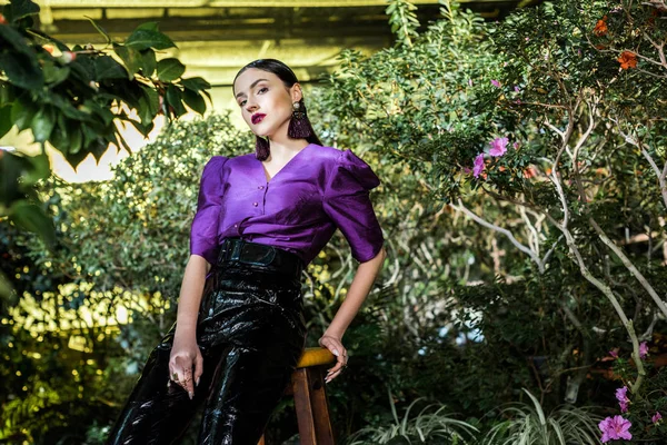 Sensual young woman in leather pants sitting on wooden chair in orangery