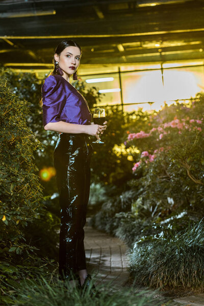 Stunning young woman in purple blouse holding wine glass in orangery