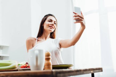 Gülümseyen kız akıllı telefon tutarak ve mutfakta selfie alarak düşük açı görünümü