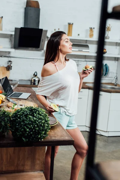 Pretty stylish girl holding cut avocado near table in kitchen