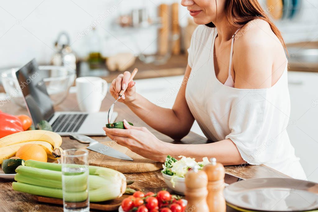 Partial view of woman eating avocado with spoon in kitchen