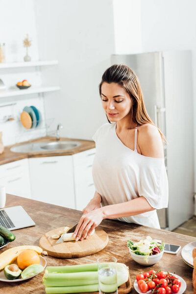 Beautiful young woman cutting banana with knife in kitchen