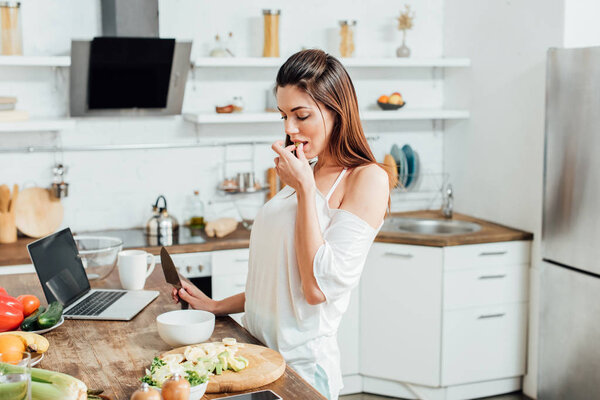 Young woman with knife cooking salad at table in kitchen