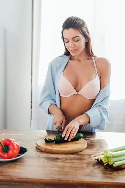 性感的女人在胸罩和衬衫切割黄瓜与刀在厨房 — 图库照片