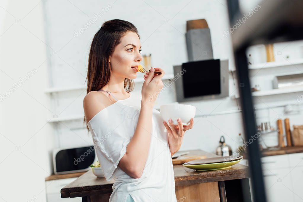 Dreamy woman holding bowl and eating cut fruit in kitchen