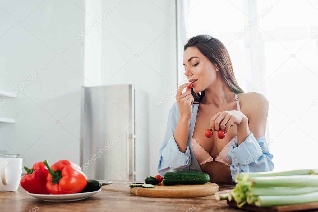 Sexy girl in bra and shirt eating cherry tomatoes in kitchen