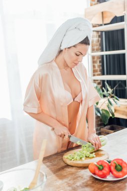 Sexy woman in housecoat with towel on head cutting celery with knife in kitchen clipart
