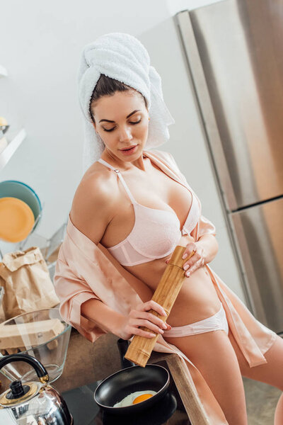Sexy young woman in lingerie with towel on head cooking fried egg in kitchen