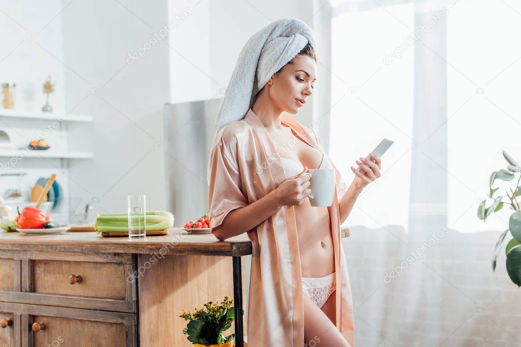 Sexy girl in lingerie and housecoat with towel on head holding cup of coffee and using smartphone in kitchen