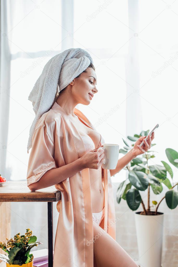 Sexy girl in lingerie and housecoat with towel on head holding cup of coffee and using smartphone in kitchen