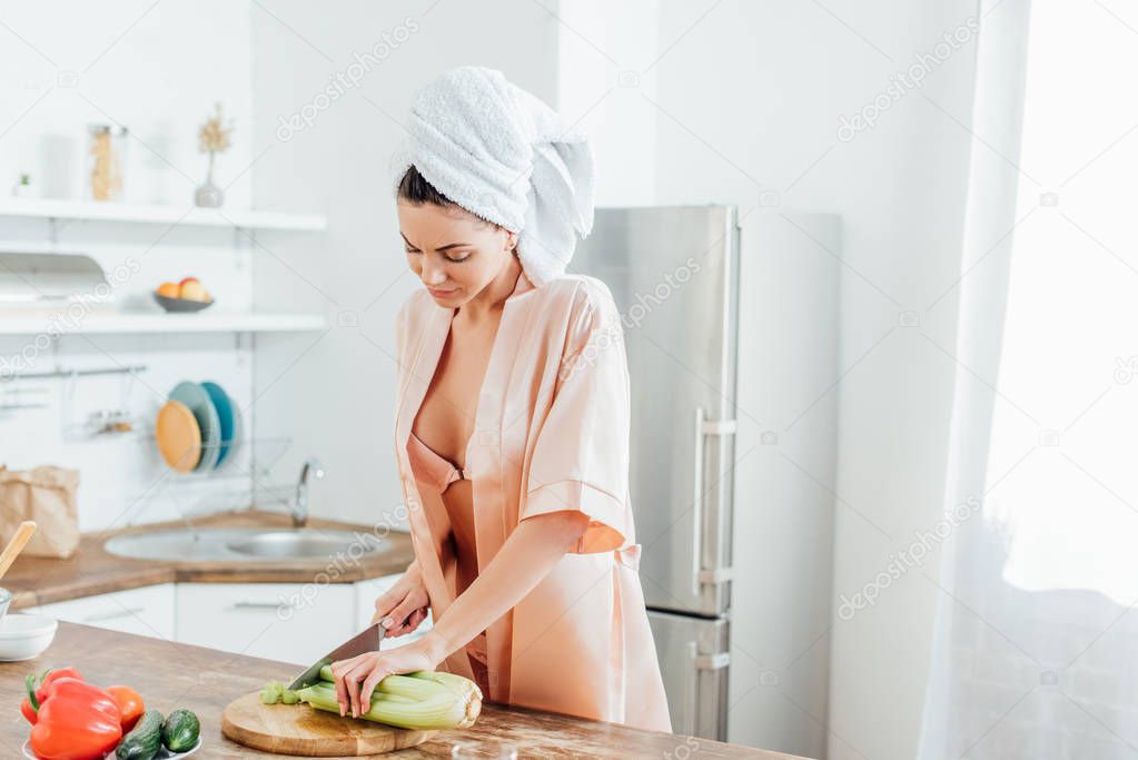 Sexy woman in housecoat with towel on head cutting celery with knife in kitchen