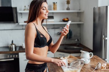 Siyah iç çamaşırı kadın fırçalamak ile yumurta kırbaçlanan ve mutfakta akıllı telefon tutarak