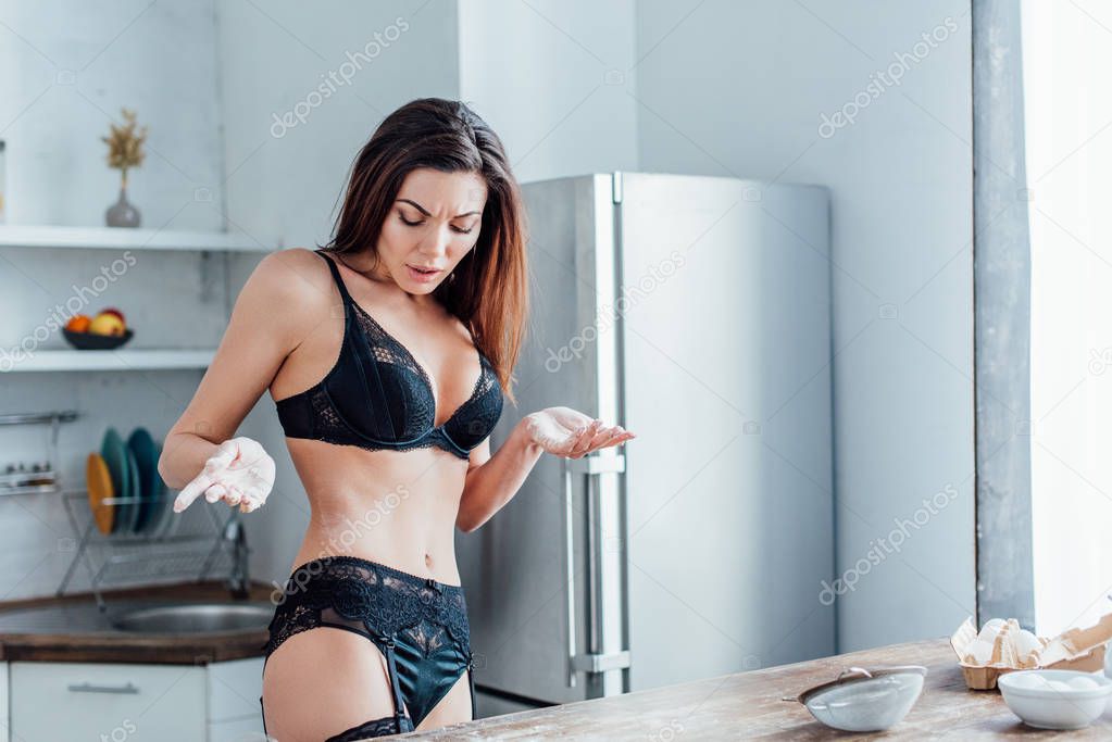 Upset woman in black lingerie standing near wooden table in kitchen