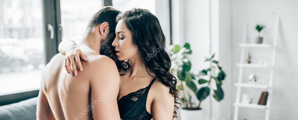 panoramic shot of attractive brunette woman with closed eyes hugging of shirtless man in bedroom 