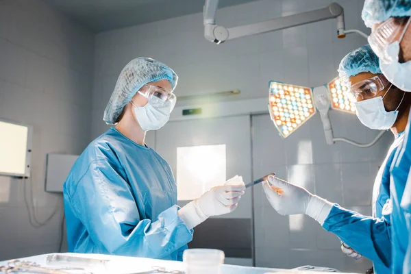 nurse in uniform and medical cap giving scalpel to doctor in operating room