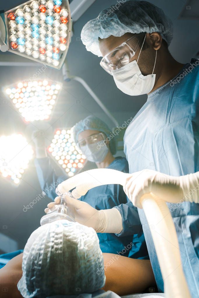 selective focus of surgeon in uniform and goggles putting mask on patient 