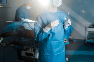 cropped view of nurse in uniform and medical mask holding goggles in operating room clipart