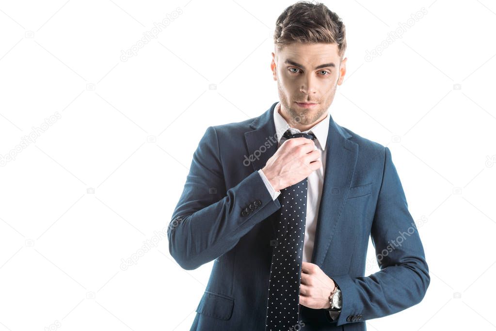 handsome, confident businessman in suite touching tie isolated on white