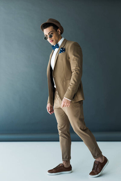 stylish mixed race man in suit, hat and sunglasses posing on grey