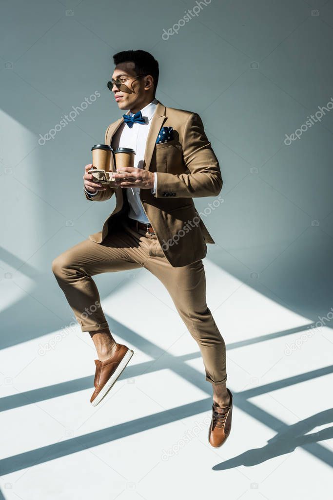 stylish mixed race man in suit holding coffee to go and jumping on grey with sunlight