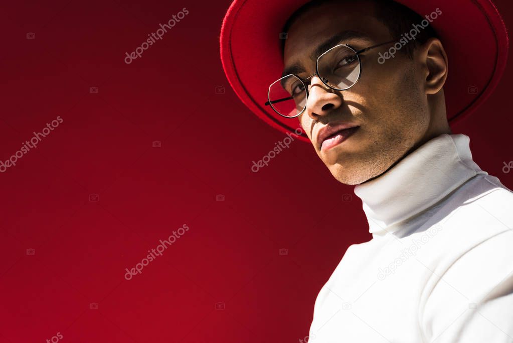 stylish mixed race man in hat and glasses looking at camera while posing isolated on red with copy space