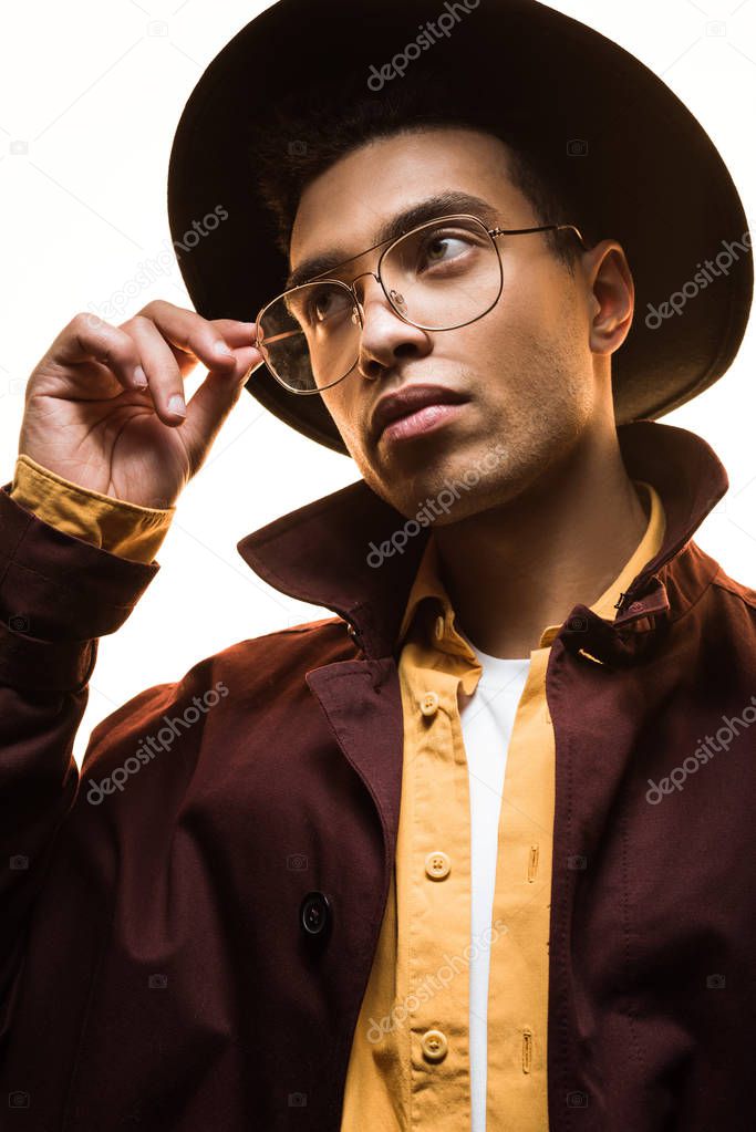 stylish mixed race man in hat holding glasses and posing isolated on white