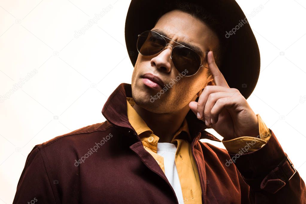 pensive stylish mixed race man in hat and sunglasses gesturing isolated on white