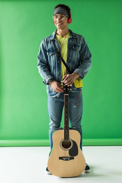 英俊的混血男子在粗斜纹棉布微笑 并提出与声学吉他绿色 — 图库照片