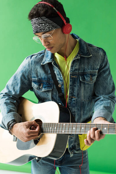 Красивый смешанный гоночный мужчина в джинсах и наушниках, играющий на акустической гитаре на зеленом
