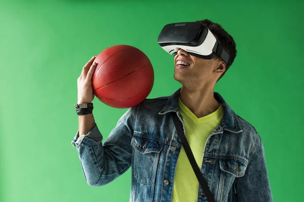 mixed race man in virtual reality headset holding basketball and smiling on green screen
