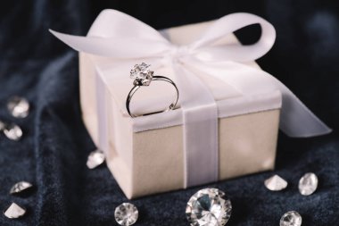 selective focus of engagement ring on gift box with bow near shiny diamonds on blue cloth clipart