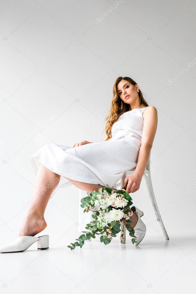 low angle view of young woman in dress sitting on chair and holding bouquet of flowers with eucalyptus leaves on white 