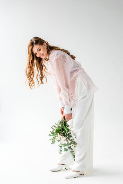 happy and stylish young woman holding flowers with eucalyptus leaves on white 