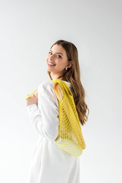 happy young woman holding yellow string bag with baguette and bottle of milk on white 