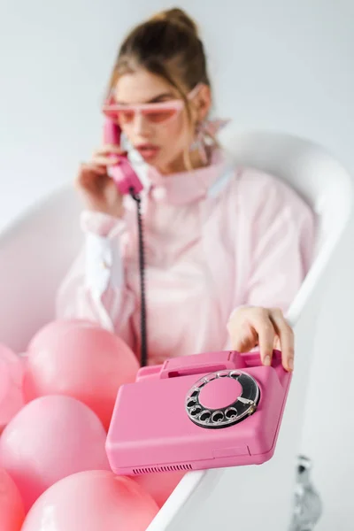 selective focus of pink retro phone near girl in sunglasses lying in bathtub with air balloons on white