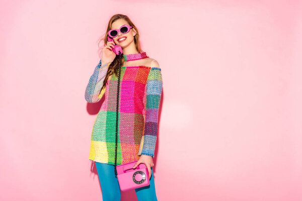 cheerful young woman in sunglasses and checkered dress talking on retro phone on pink